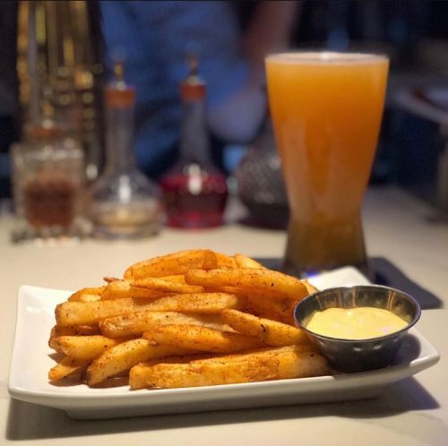 A plate of golden fries with a small bowl of dipping sauce, alongside a tall glass of orange beverage.