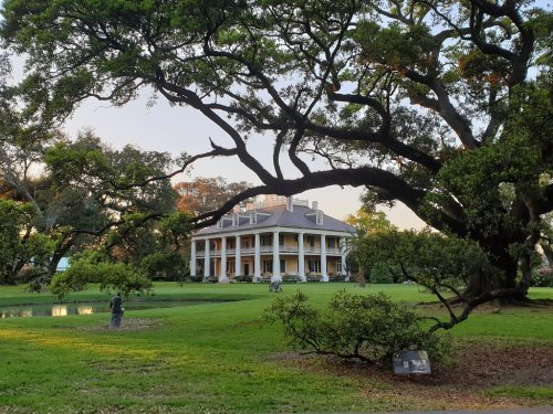 A grand mansion surrounded by lush greenery and large trees, with a serene pond in the foreground.