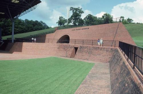 A brick structure with an archway, surrounded by green grass and trees, featuring the words "Cairo Museum" on the wall.