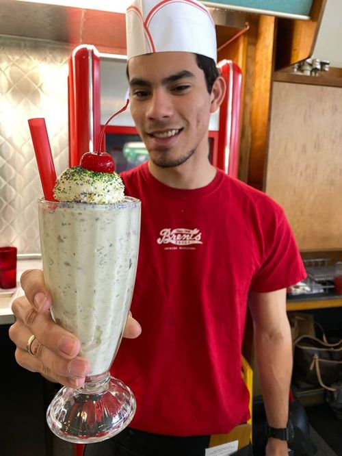 A young man in a red shirt holds a tall milkshake topped with whipped cream and a cherry, smiling at the camera.