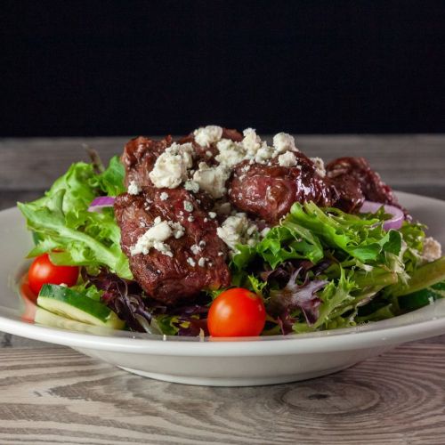 A fresh salad topped with grilled meat, crumbled cheese, cherry tomatoes, and mixed greens on a white plate.