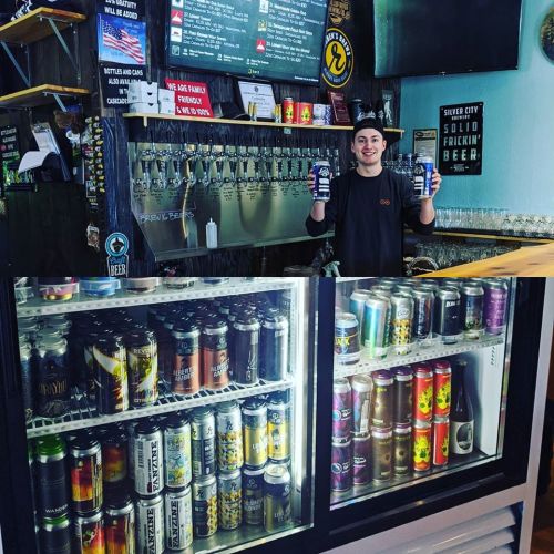 A bartender holds two drinks in front of a wall of taps and a fridge filled with various canned beers.