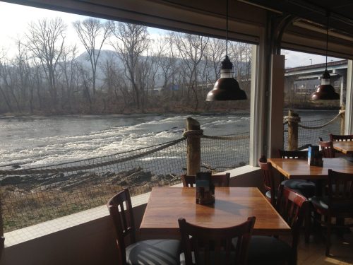 View of a river with rapids from a restaurant, featuring wooden tables and chairs in the foreground.