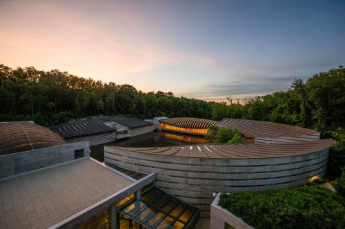 A modern architectural structure with curved roofs, surrounded by trees and reflecting in a calm body of water at sunset.