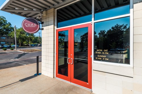 Red double doors of Char Restaurant, with a sign displaying operating hours, set against a sunny outdoor backdrop.