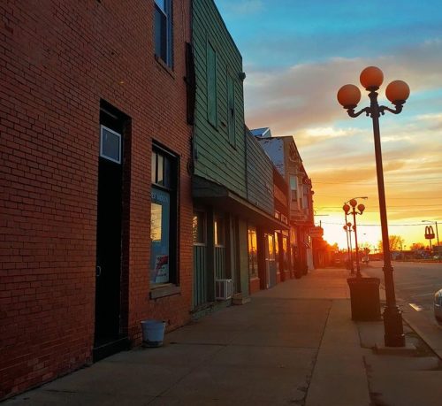 Sunset casts warm light on a quiet street lined with brick buildings and vintage street lamps.
