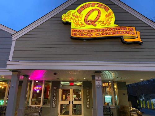 Exterior of Gotta Q Smokehouse & BBQ Catering, featuring a lit sign and welcoming entrance at dusk.