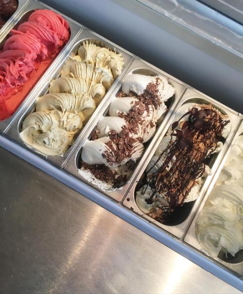 A display of various gelato flavors in metal trays, including red, white, and chocolate-topped varieties.