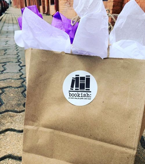 A brown paper bag with a "bookish" logo, filled with colorful tissue paper, sits on a cobblestone street.