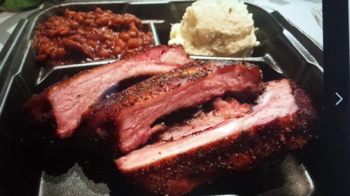 A takeout container with three smoked rib pieces, baked beans, and a scoop of mashed potatoes.