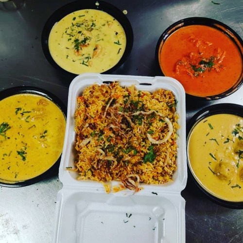 A takeout container of biryani surrounded by three bowls of curry: yellow, orange, and creamy white sauces.