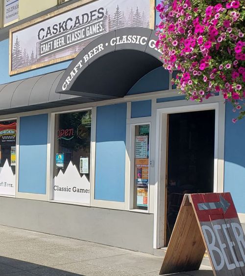 Exterior of Caskcades, a craft beer and classic games shop, featuring a colorful flower basket and an open sign.