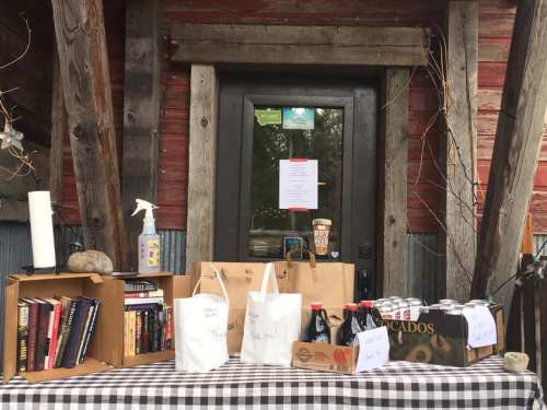 A table with books, bags, and drinks outside a rustic door, featuring a sign and a spray bottle.