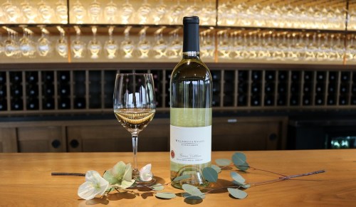 A bottle of white wine with a glass, surrounded by flowers and eucalyptus, set against a backdrop of wine glasses.