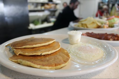 Three golden pancakes on a plate with a side of creamy white sauce, set in a busy diner environment.