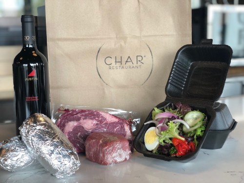 A takeout bag from CHAR Restaurant with a bottle of wine, two foil-wrapped items, a steak, and a salad in a container.
