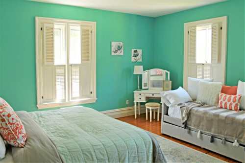 Bright turquoise bedroom with a bed, a cozy seating area, and natural light from two windows. Decorated with soft textiles.