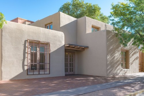 A modern adobe-style building with large windows and a wooden awning, surrounded by trees and a clear blue sky.