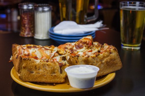 A plate of stuffed crust pizza with pepperoni and a side of ranch dressing, accompanied by drinks in the background.