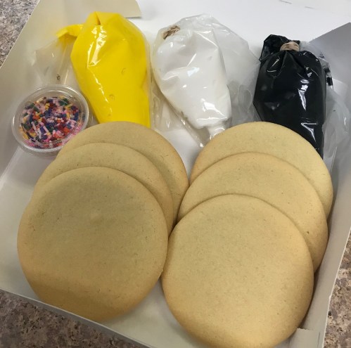 A box containing several uniced cookies, three bags of icing in yellow, white, and black, and a container of sprinkles.