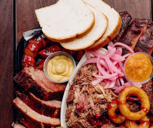 A platter of assorted meats, including pulled pork, ribs, sausage, and sides of bread, pickled onions, and sauces.