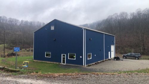 A blue metal building in a rural area, surrounded by trees and a gravel driveway, with a cloudy sky above.