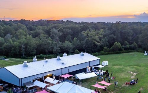 Aerial view of a venue with tents and people gathered outdoors at sunset, surrounded by trees and open fields.