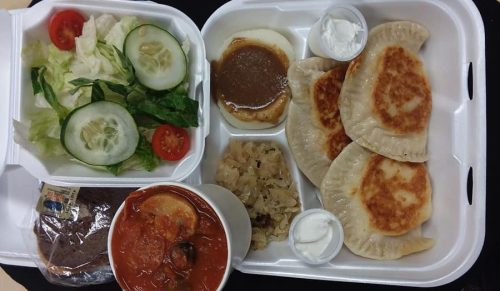 A takeout container with three fried dumplings, mashed potatoes, salad, a cup of soup, and a dessert.