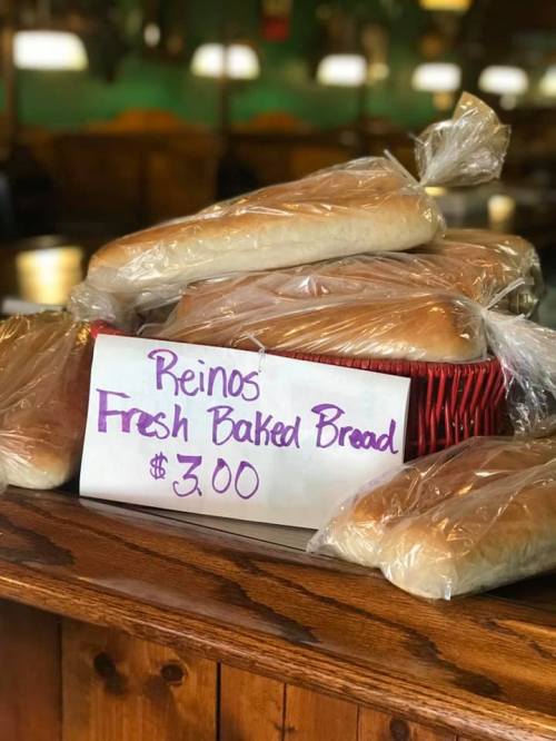 A basket of fresh baked bread loaves wrapped in plastic, with a sign reading "Reinos Fresh Baked Bread $3.00."