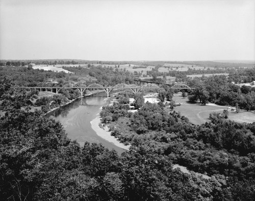 Aerial view of a river winding through lush greenery, with a large arch bridge spanning across it.