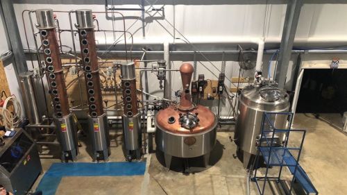 A distillery setup featuring copper and stainless steel equipment, including columns and a large fermentation tank.