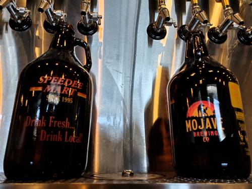 Two glass growlers on a stainless steel counter, one labeled "Speedee Mart" and the other "Mojave Brewing Co."