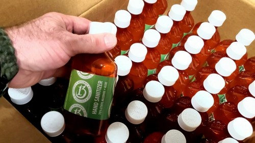 A hand holding a bottle of hand sanitizer above a box filled with multiple sanitizer bottles.