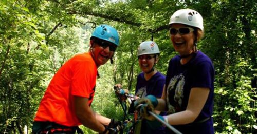 Three people in helmets and harnesses smile while zip-lining through a lush green forest.
