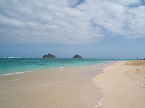A serene beach with soft sand and turquoise waters, featuring two small islands in the distance under a partly cloudy sky.