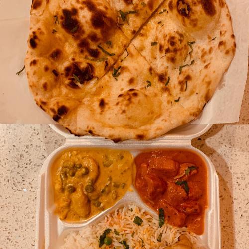 A takeout container with rice, two curries, and a piece of naan bread on top, garnished with herbs.