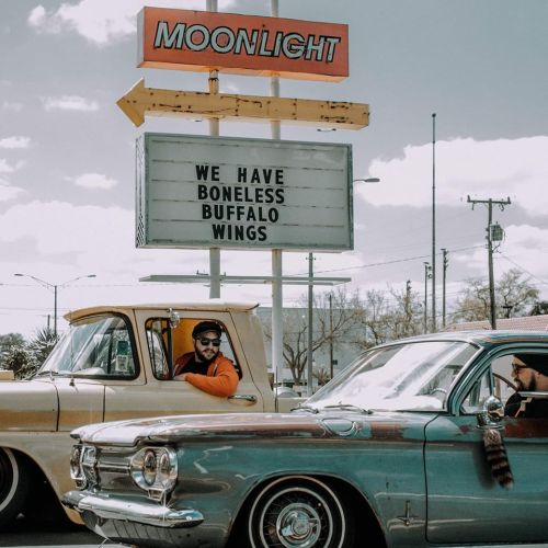 A vintage car show with two classic cars parked near a sign advertising boneless buffalo wings at Moonlight.