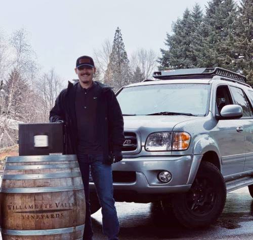 A man stands next to a silver SUV and a wine barrel, holding a box, with trees in the background.
