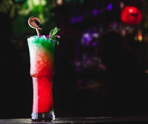 A colorful layered cocktail in a tall glass, garnished with mint, set against a dimly lit bar background.