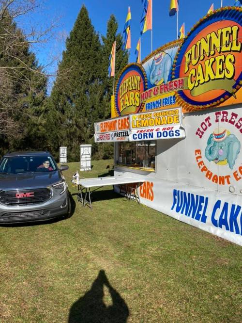 A food truck offering elephant ears, funnel cakes, and corn dogs, parked on grass with trees in the background.