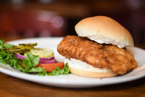 A crispy fried chicken sandwich on a bun, served with lettuce, tomato, pickles, and onion on a plate.