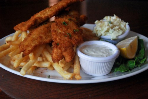 A plate of crispy fried fish fillets served with French fries, coleslaw, tartar sauce, and a lemon wedge.