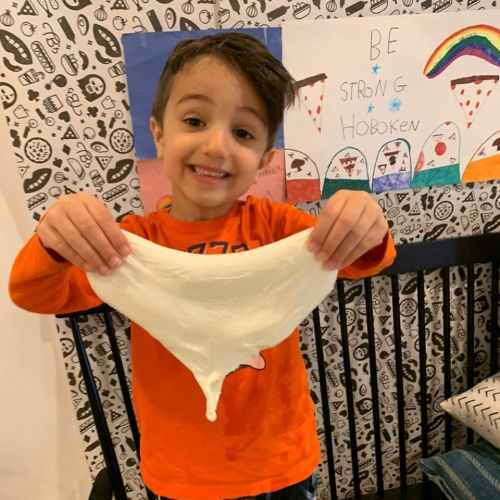 A smiling child in an orange shirt holds a stretchy piece of slime, with colorful drawings in the background.