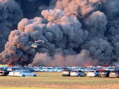 A helicopter hovers over a field filled with cars, engulfed in thick black smoke and flames in the background.