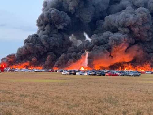 A large fire produces thick black smoke and flames, with cars visible in the foreground and a field in the background.