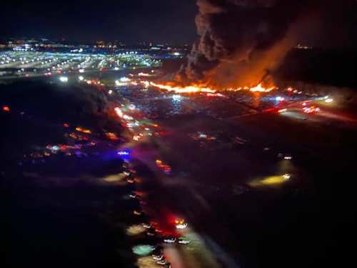 A large fire burns at night, with flames and smoke rising above a field, while emergency vehicles respond nearby.