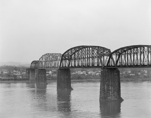Black and white image of a large, arched bridge spanning a river, with stone pillars and a cloudy sky in the background.