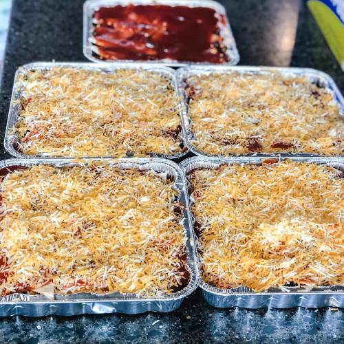 Four aluminum trays filled with layered pasta, sauce, and a golden cheese topping, ready for baking.