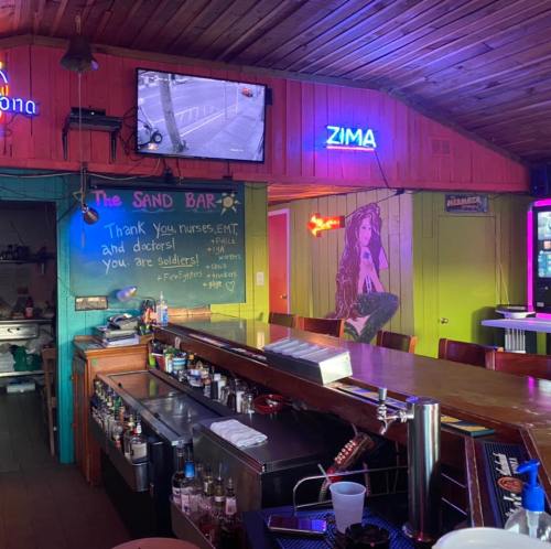 A colorful bar interior with a chalkboard message thanking healthcare workers, a TV, and neon signs.