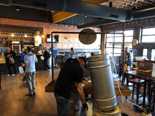 A brewery setup with a person working on equipment, while others are seen in the background, inside a rustic bar.